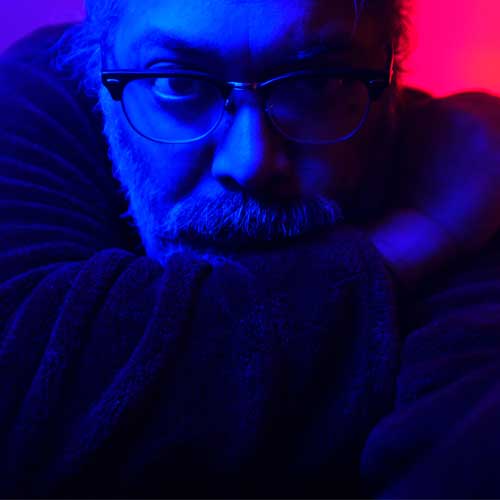 Portrait of Arnie Guha bathed in blue lighting with a bright pink light visible behind him