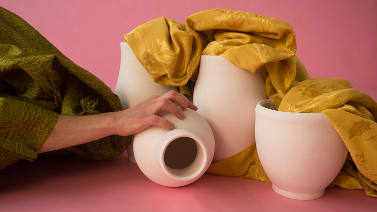 A hand grasping a white ceramic vessel and draped fabric