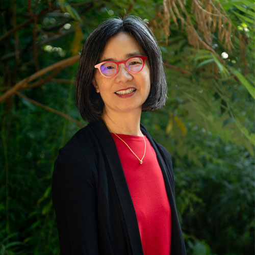 A photographic portrait of Catherine Little wearing red classes, a red shirt, and a black cardigan. She is standing in front of some greenery.
