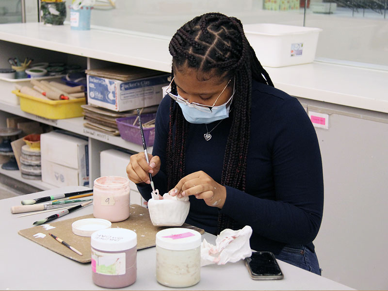 A woman with long braids wearing a mask and glazing pottery