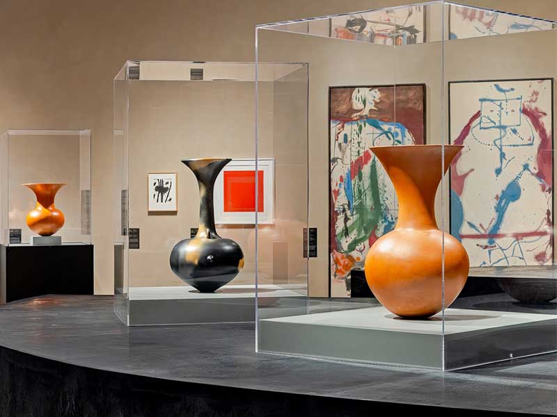 Orange and black ceramic vessels in glass vitrines with abstract paintings visible behind them
