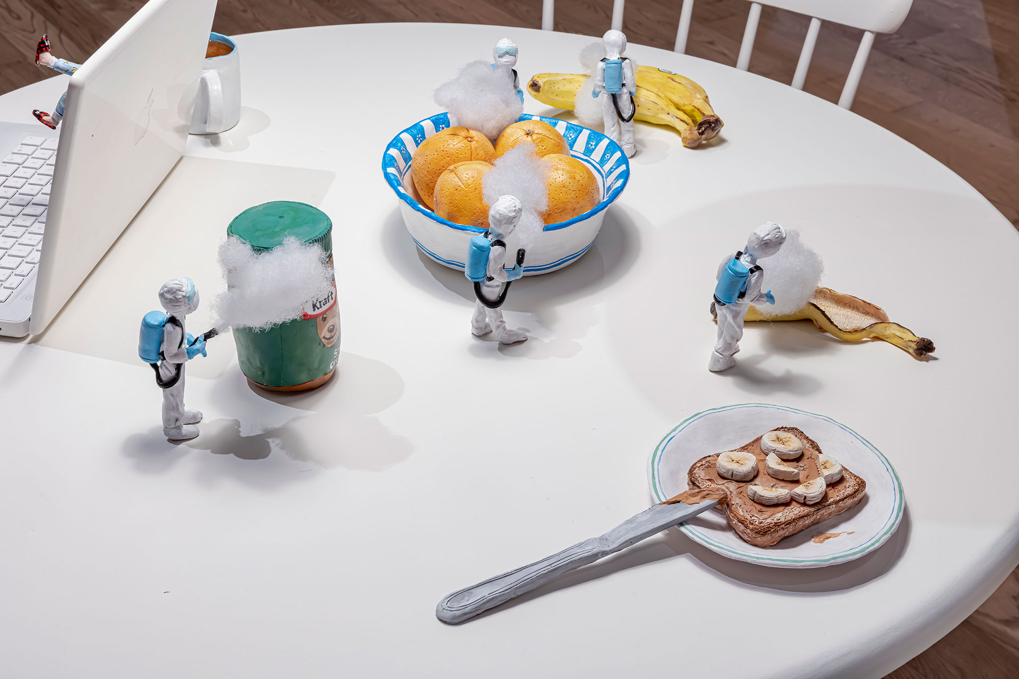 A white kitchen table with polymer clay food and miniature polymer clay figures in hazmat suits spraying the food