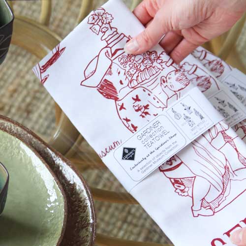 A hand holding a white tea towel with a red illustration of a ceramic figure