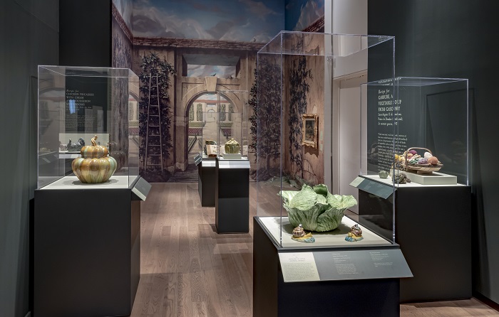 Savour: Food Culture in the Age of Enlightenment. A painted garden archway is in the background. Cases filled with ceramic objects including a cabbage, pumpkin, as well as knitted vegetables are in the foreground