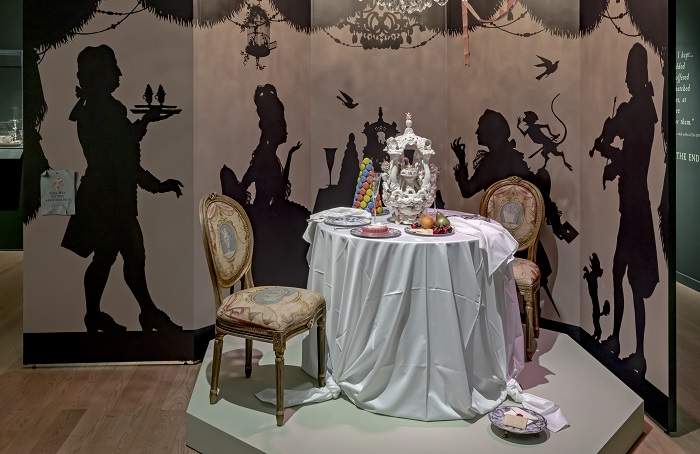A dining table with a ceramic centrepiece, and fake food. A painted backdrop of a dining scene is in the background