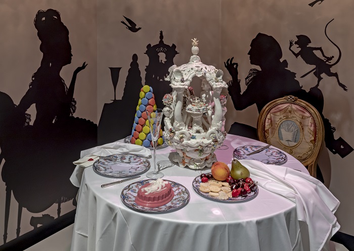 A dining table with plates of realistic ceramic and plastic food, and a painted backdrop of a romantic dining scene is in the background