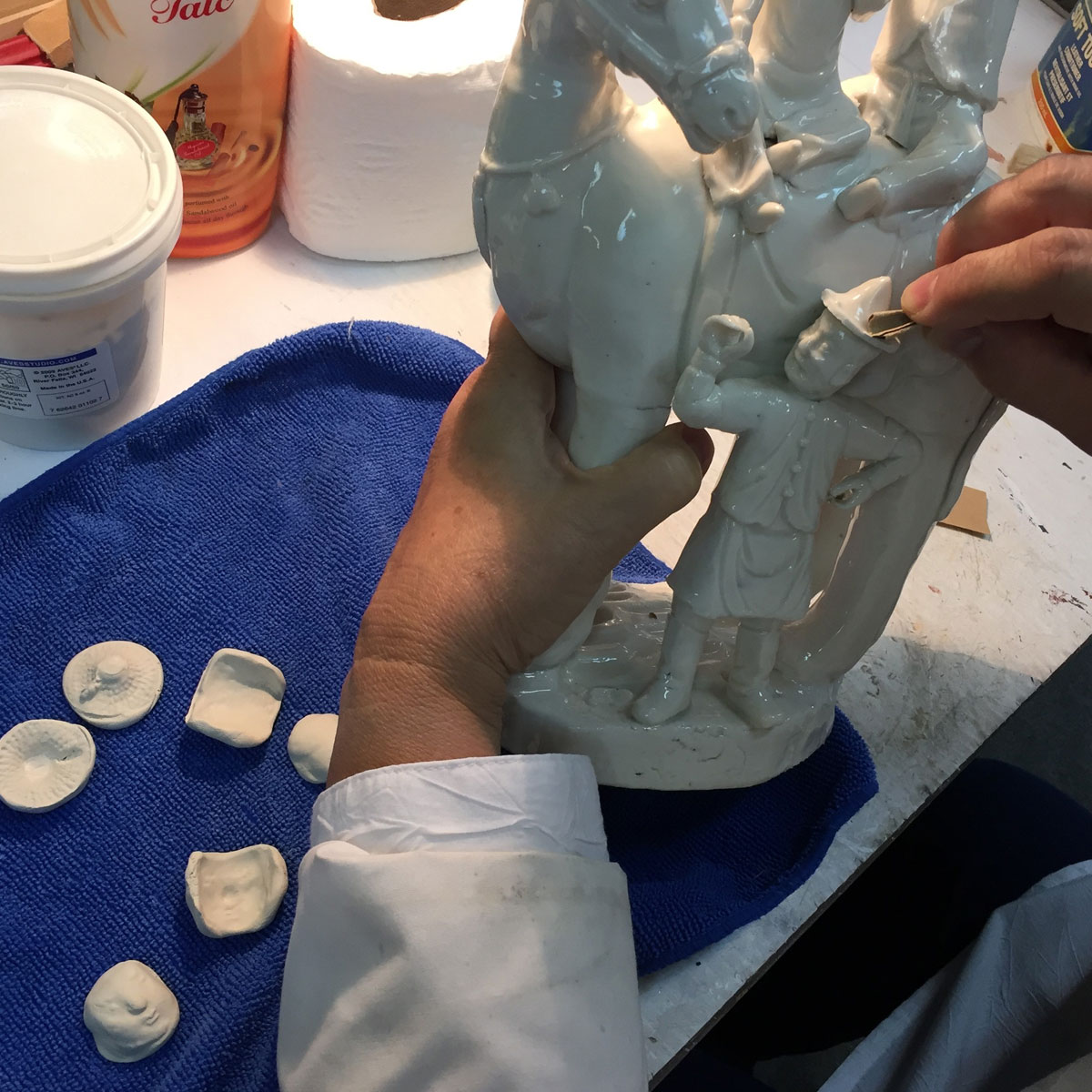 Hands using glass paper to shape the clay hat of a white porcelain figure group