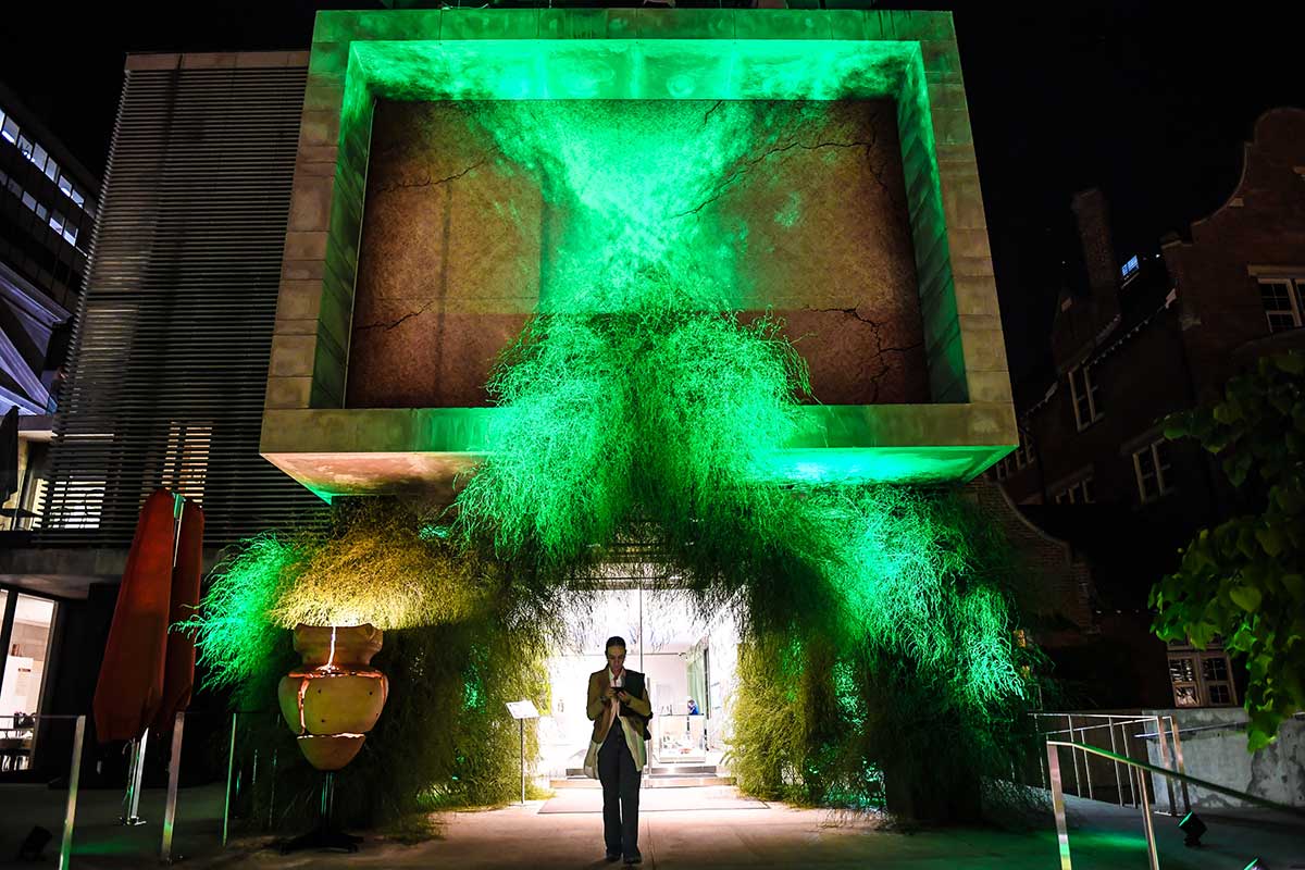 The exterior of the Gardiner Museum with a floral installation around the entrance and a large green X projected on the facade