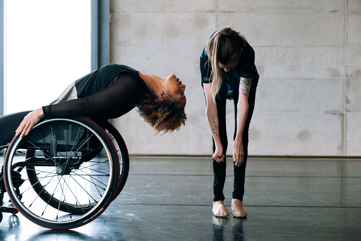 A dancer who uses a wheelchair leaning back toward another dancer who is reaching toward their toes