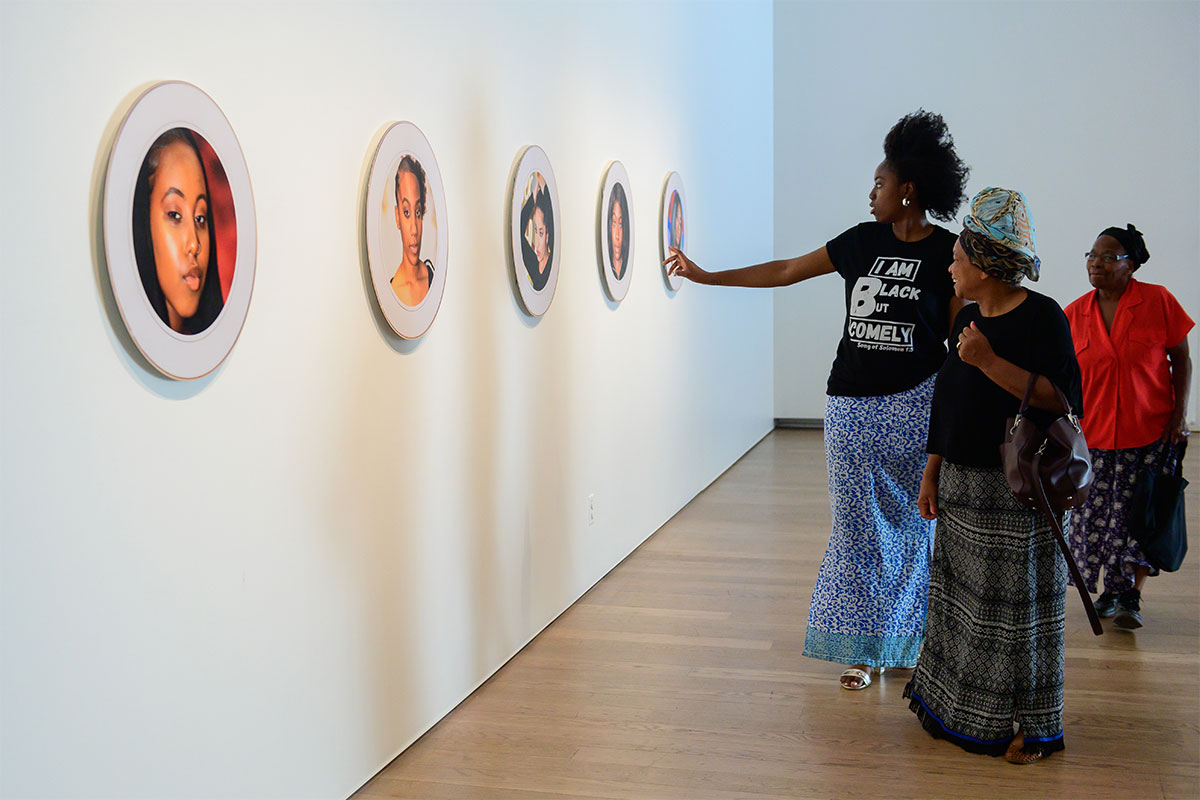 People in the gallery looking at a series of round portraits of young racialized women on the wall