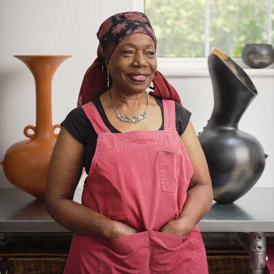 Artist Magdalene Odundo in her studio wearing a headscarf and red overalls smiling between two ceramic vessels