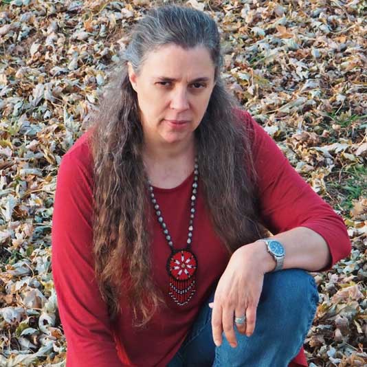 Portrait of Melanie McKay-Cody kneeling in a red shirt and beaded necklace