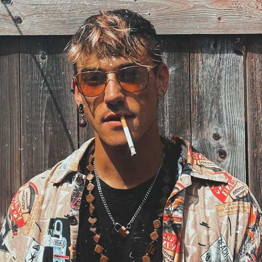 A white man with blond hair wearing sunglasses with a cigarette in his mouth