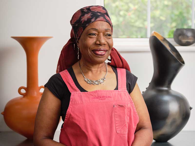 Magdalene Odundo in her studio standing between two vessels and wearing a red apron and headscarf