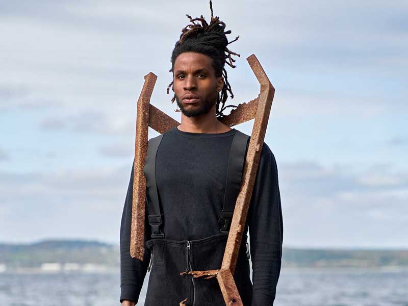 Artist Oluseye wearing black overalls and a wood structure in front of a seascape