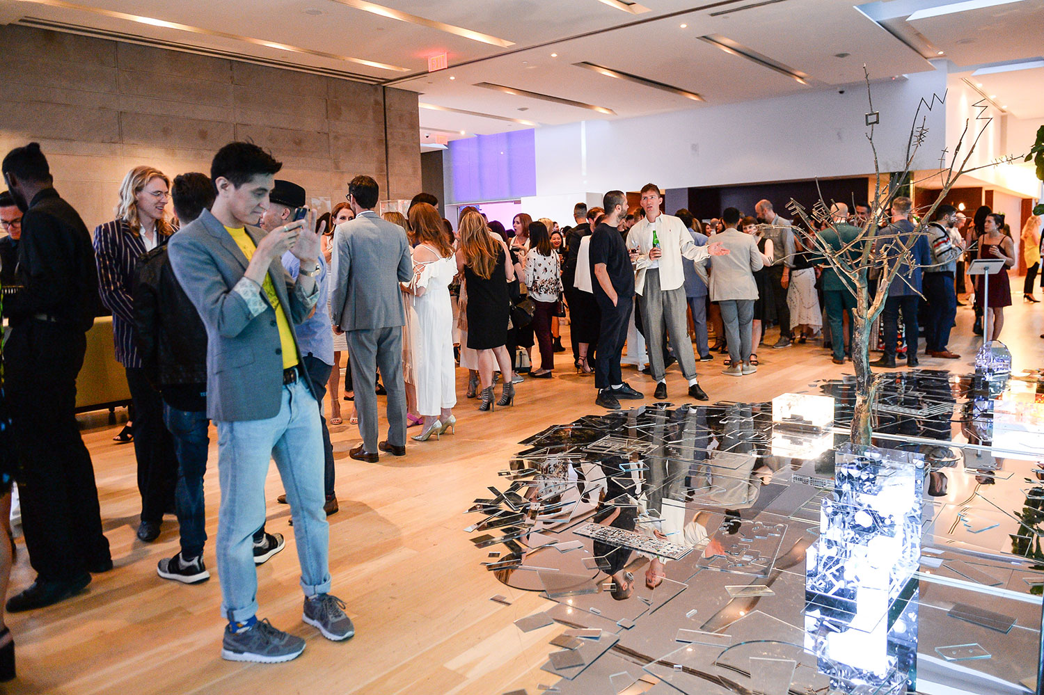 A crowded room of people with a man in the foreground taking a photo of an art installation made up of broken mirror shards on the floor and a tree