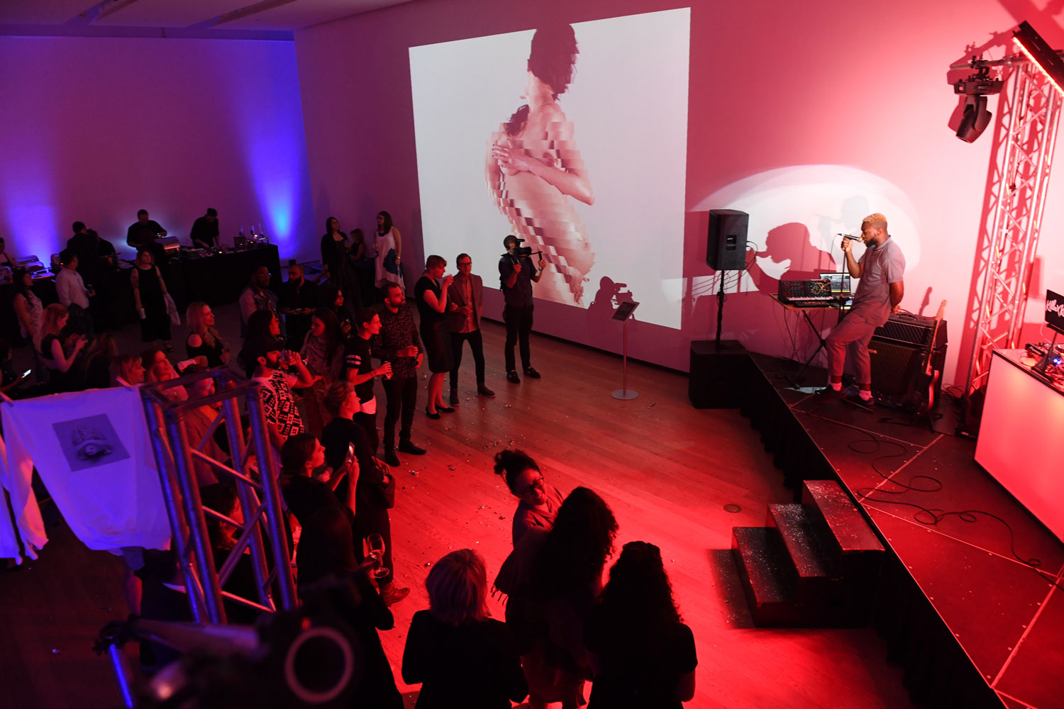 Room lit in red and purple with people watching a singer perform. There is a digital projection of a human figure on the wall.