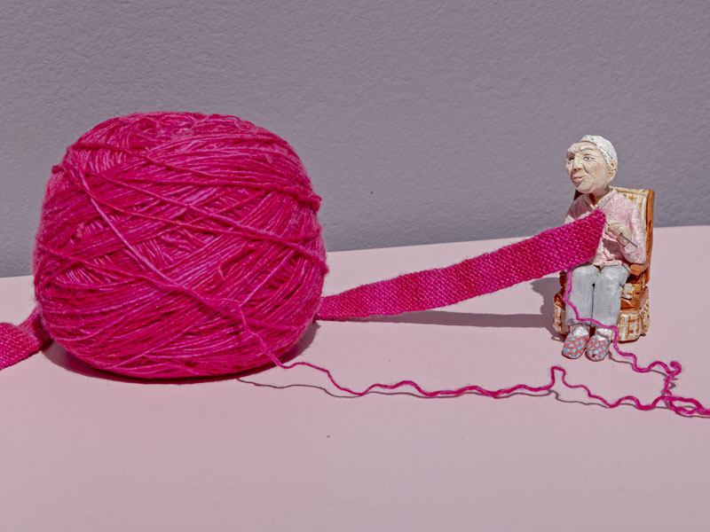 An miniature clay figure of an elderly woman in a rocking chair knitting from a full-size ball of yarn