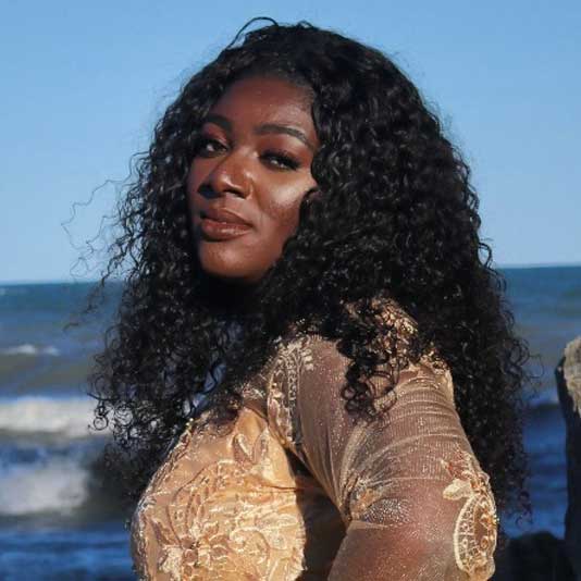 Tracey Kayy wearing a gold dress in front of the ocean
