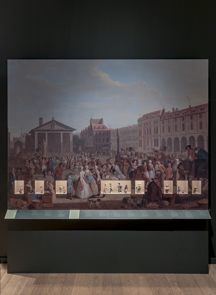 A painted scene of a bustling 18th century marketplace. Ceramic figurines are inserted in cutouts in the painting