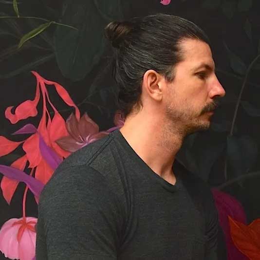 Profile portrait of artist Zachari Logan in a black t-shirt with his hair in a bun, seated in front of a painted mural